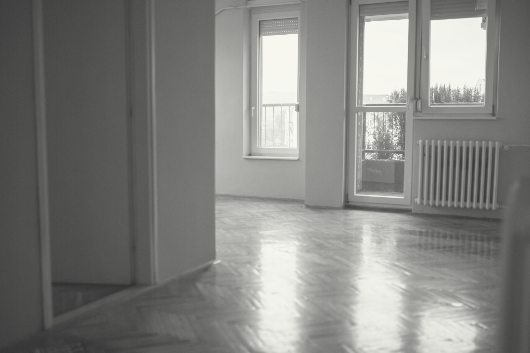 An empty apartment. The photo is taken from the entrance of a room with a view of the apartment’s balcony and two windows. The hardwood floors and walls are bare.