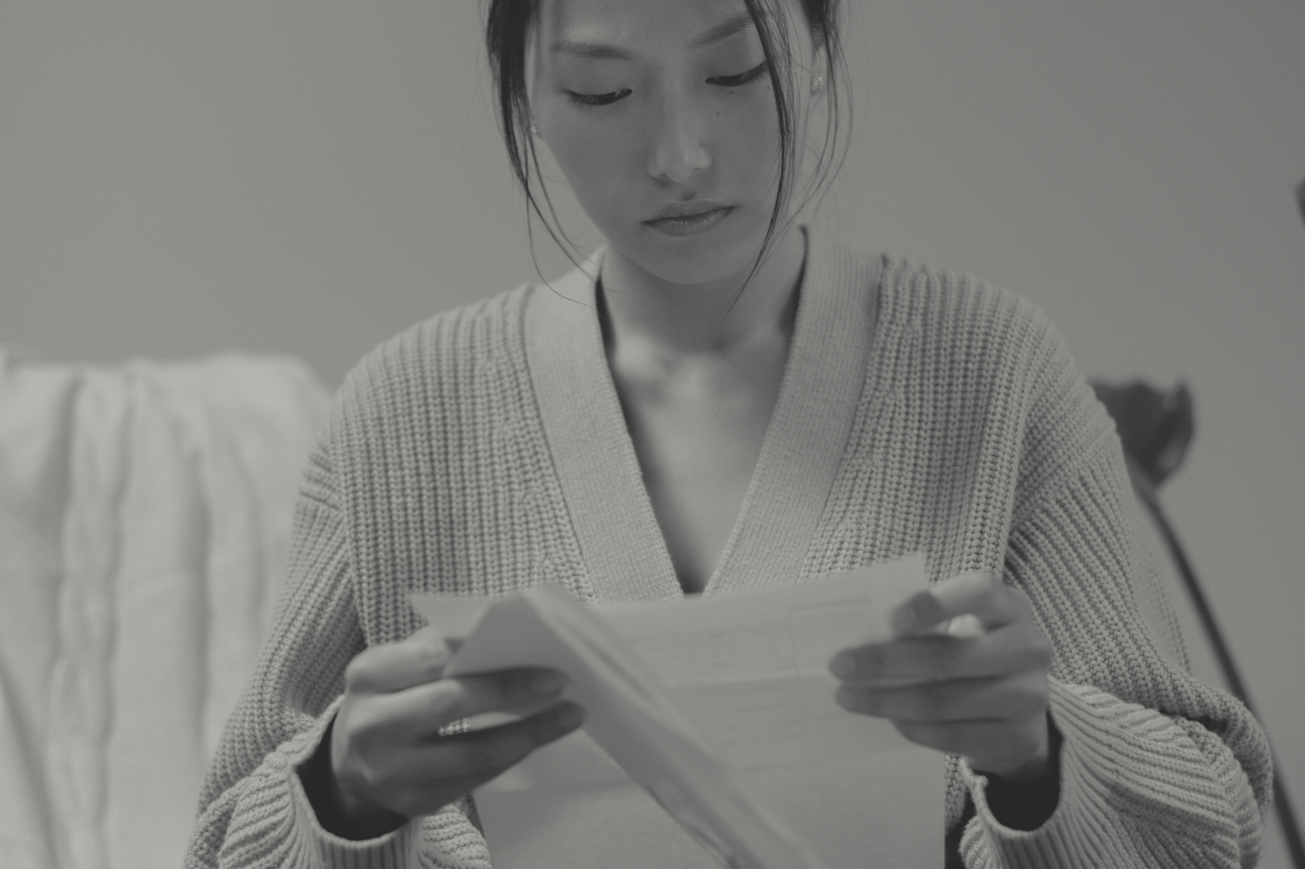 A woman is sitting and reading a letter. She has a fixed expression and is looking down at the letter in her hands.